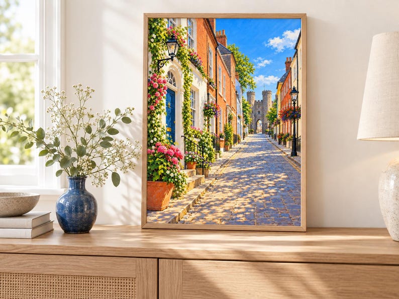 May include: A framed wall art print depicting a cobblestone street lined with colorful brick buildings, climbing green vines, and pink flowers. A blue door is visible on the left, and a stone archway stands in the distance under a bright blue sky.