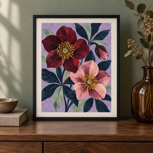 K&ouml;nnte beinhalten: Ein gerahmter Kunstdruck mit einer botanischen Illustration von zwei Nieswurzbl&uuml;ten, eine dunkelrot und eine zartrosa, mit gelben Mitten und dunkelgr&uuml;nem Blattwerk vor einem strukturierten hellvioletten Hintergrund, in einem schwarzen Rahmen.