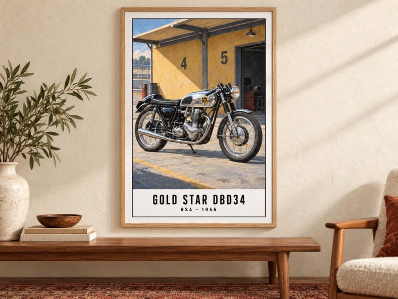 May include: A framed wall art print featuring a 1956 BSA Gold Star DBD34 motorcycle parked in front of a yellow garage building with the numbers 4 and 5 visible. The text at the bottom reads GOLD STAR DBD34 BSA 1956 in a clean black font.
