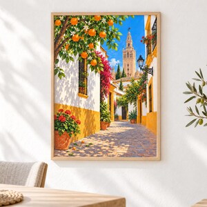 May include: A framed wall art print depicting a sunlit Mediterranean street scene with white buildings featuring yellow trim, vibrant orange trees, pink bougainvillea, terracotta pots with red flowers, and a tall stone bell tower under a blue sky.