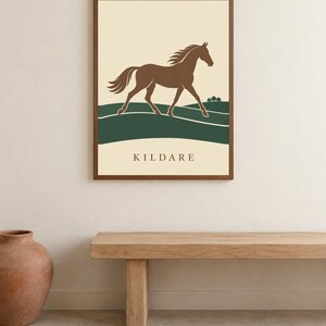 May include: A framed wall art print featuring a brown horse galloping across green rolling hills against a cream background. The word KILDARE is printed in dark brown capital letters at the bottom center of the rectangular print.