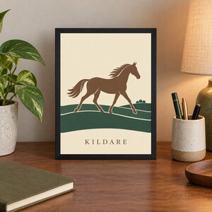May include: A framed rectangular print featuring a brown horse silhouette galloping across a dark green landscape against a cream background. The word KILDARE is printed in a simple serif font at the bottom center of the artwork within the frame.