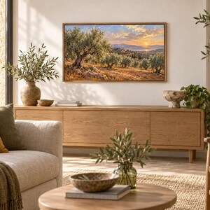 May include: A rectangular landscape painting in a thin wooden frame depicting an olive grove at sunset with rolling hills and a distant body of water under a soft orange and blue sky, mounted on a light-coloured wall above a wooden media console.