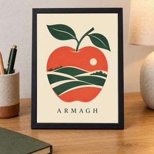 May include: A framed print featuring a stylized red apple illustration on a cream background. The apple contains a landscape of green rolling hills and a small white sun. The word ARMAGH is printed in dark green capital letters at the bottom.