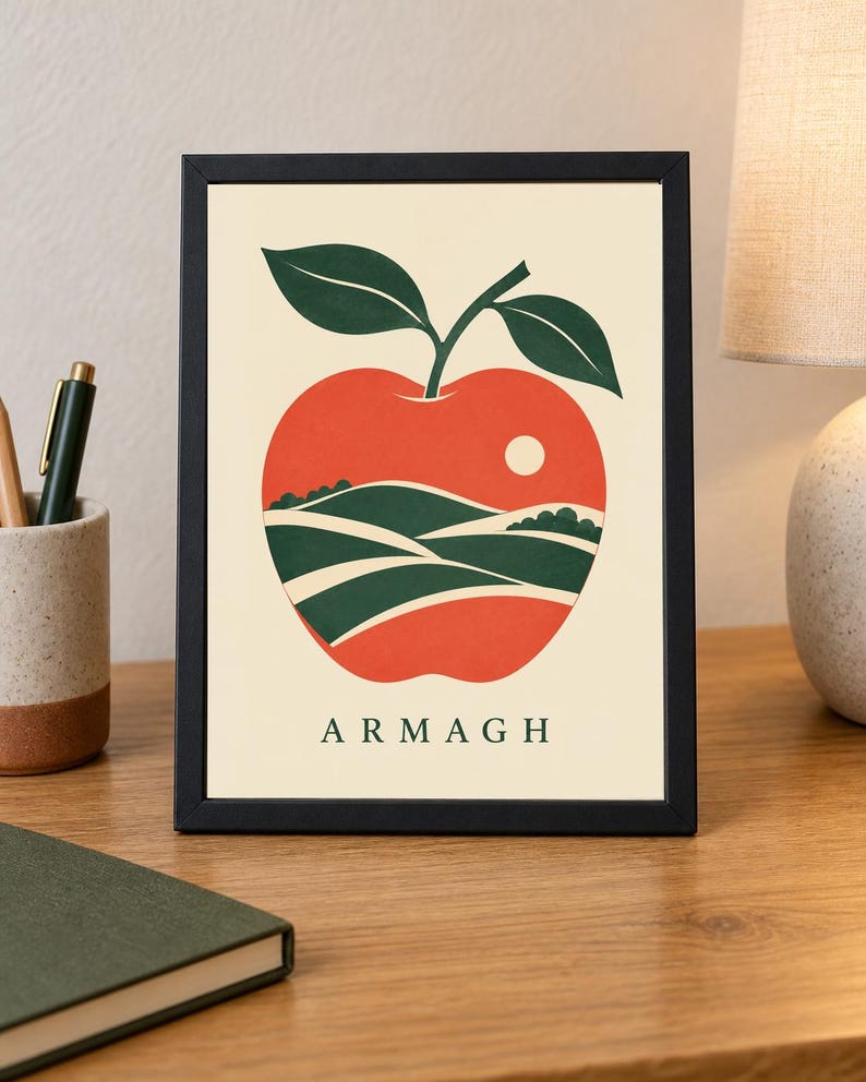 May include: A framed print featuring a stylized red apple illustration on a cream background. The apple contains a landscape of green rolling hills and a small white sun. The word ARMAGH is printed in dark green capital letters at the bottom.