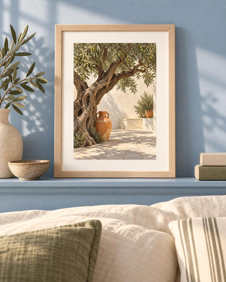 May include: A framed wall art print featuring a gnarled olive tree with green leaves, a terracotta amphora, and a stone wall in a sunlit courtyard. The print is housed in a light-toned wooden frame with a white mat, displayed on a blue shelf.