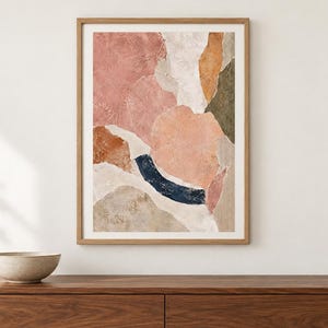 May include: A framed rectangular wall art print featuring an abstract composition of layered organic shapes in shades of dusty pink, terracotta, cream, olive green, and a deep navy blue curved brushstroke, all enclosed in a light wood frame.