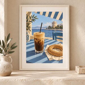 May include: A framed art print depicting a glass of iced coffee with a black straw and a sesame seed bagel on a blue table. The scene features a coastal view with a white tower, blue sea, and striped awning, rendered in a painterly style.