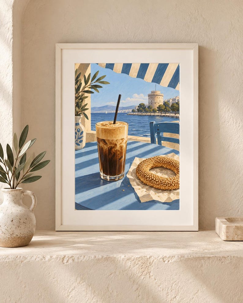 May include: A framed art print depicting a glass of iced coffee with a black straw and a sesame seed bagel on a blue table. The scene features a coastal view with a white tower, blue sea, and striped awning, rendered in a painterly style.