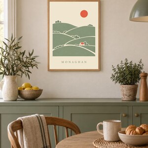 May include: A framed wall art print featuring a stylised landscape with rolling green hills, a small red house, and a red sun against a cream background. The word MONAGHAN is printed in black capital letters at the bottom of the illustration.