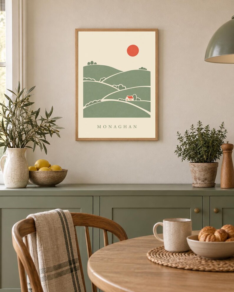 May include: A framed wall art print featuring a stylised landscape with rolling green hills, a small red house, and a red sun against a cream background. The word MONAGHAN is printed in black capital letters at the bottom of the illustration.