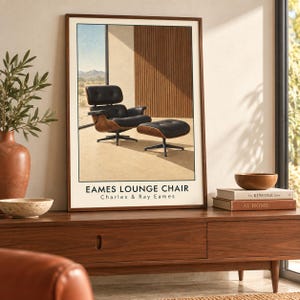 May include: A framed wall art print featuring a black leather Eames lounge chair and matching ottoman on a light floor. The text EAMES LOUNGE CHAIR Charles & Ray Eames is printed at the bottom. The background shows a desert landscape and wood paneling.