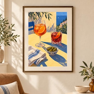 May include: A framed art print depicting a yellow table with an orange spritz in a stemmed glass, a dark red cocktail in a tumbler, and a small bowl of green olives. A white napkin with blue stripes sits nearby, set against a coastal Mediterranean backdrop.