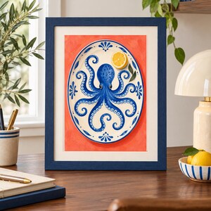 May include: A framed art print featuring an oval white platter with a blue octopus illustration and a single lemon slice. The platter is centred against a solid coral-orange background. The entire image is enclosed in a dark blue wooden frame.