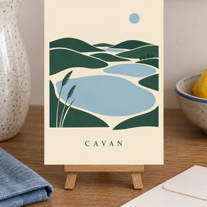 May include: A rectangular art print on a small wooden easel featuring a stylized landscape illustration of green rolling hills, light blue bodies of water, and a pale blue sun against a cream background with the word CAVAN printed in black.