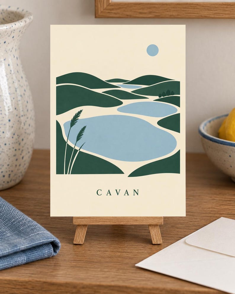 May include: A rectangular art print on a small wooden easel featuring a stylized landscape illustration of green rolling hills, light blue bodies of water, and a pale blue sun against a cream background with the word CAVAN printed in black.