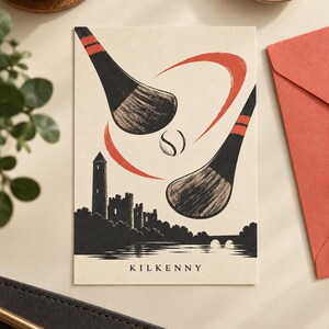 Peut inclure: Une carte de v&oelig;ux rectangulaire sur fond cr&egrave;me avec une illustration noire et orange de deux crosses de hurling et une balle au-dessus de la silhouette d'un ch&acirc;teau et d'une rivi&egrave;re. Le mot KILKENNY est imprim&eacute; en noir en bas au centre.