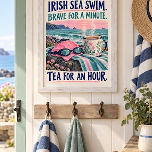 May include: A framed wall art print featuring a coastal scene with a pink swim cap, goggles, and a floral mug. The text reads: IRISH SEA SWIM. BRAVE FOR A MINUTE. TEA FOR AN HOUR. The illustration includes a sunset over the ocean and rocky cliffs.