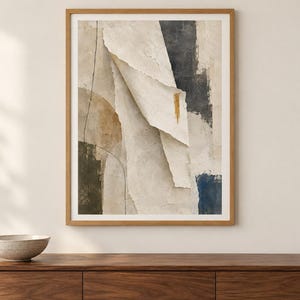 May include: A framed abstract wall art print featuring layered paper-like textures in shades of cream, beige, and charcoal gray. The composition includes a prominent vertical fold, a thin black line, and a small gold accent mark on a textured surface.