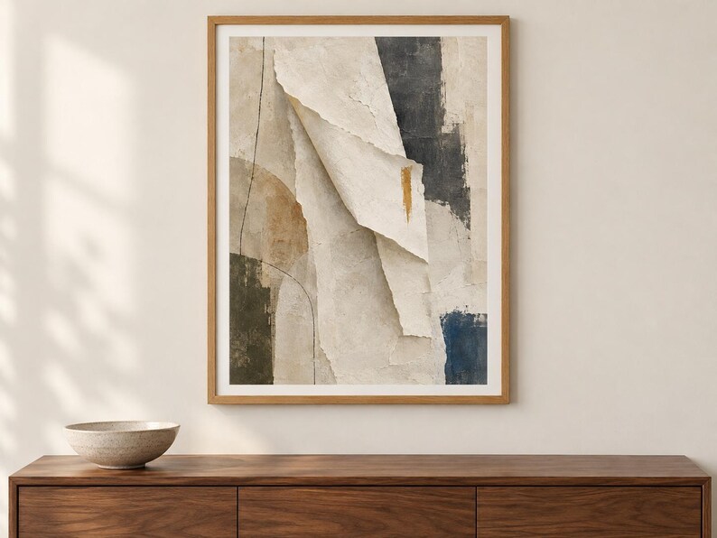 May include: A framed abstract wall art print featuring layered paper-like textures in shades of cream, beige, and charcoal gray. The composition includes a prominent vertical fold, a thin black line, and a small gold accent mark on a textured surface.