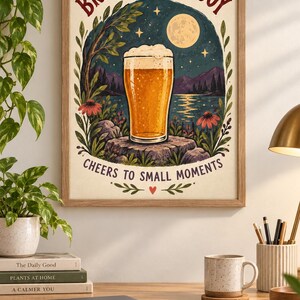 May include: A framed wall art print featuring a glass of beer on a rock, set against a night landscape with a full moon, mountains, and a lake. The text reads BREW A LITTLE JOY and CHEERS TO SMALL MOMENTS in dark red lettering on a cream background.