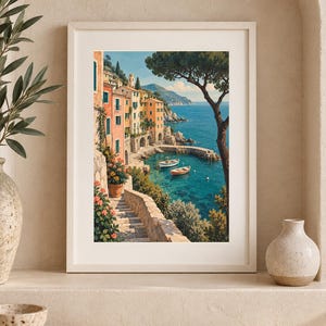 May include: A framed vertical art print depicting a coastal Mediterranean village with pastel-coloured buildings, stone stairs, and vibrant flowers leading down to a turquoise bay with two small boats floating on the water under a clear blue sky.