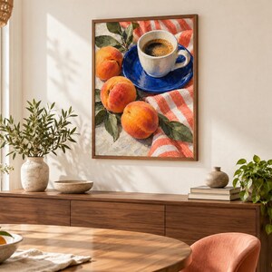 May include: A framed wall art print featuring a still life composition of three ripe orange peaches with green leaves, a white ceramic coffee cup filled with dark coffee, and a vibrant blue saucer resting on a red and white striped fabric cloth.