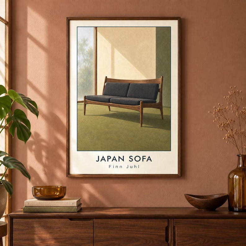 Pu&ograve; includere: Una stampa artistica da parete incorniciata che raffigura un divano in legno con cuscini grigio scuro, posizionato contro una parete chiara e un pavimento verde. Il testo JAPAN SOFA e Finn Juhl &egrave; stampato in nero sul bordo bianco del poster.