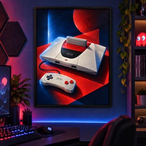May include: A framed wall art print featuring a light gray Amstrad GX4000 video game console with a cartridge inserted and a matching controller. The design includes a red and black geometric background with blue lighting accents.