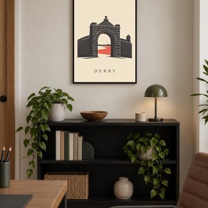 May include: A framed wall art print featuring a black graphic illustration of a stone archway and city walls with a red path underneath, set against a cream background with the text DERRY printed in black capital letters at the bottom center.