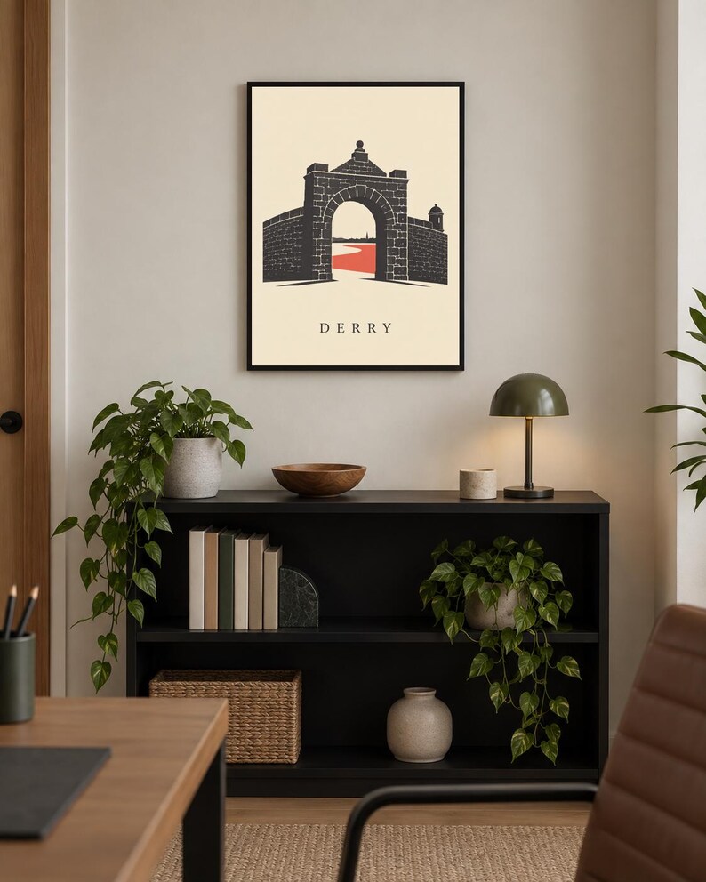 May include: A framed wall art print featuring a black graphic illustration of a stone archway and city walls with a red path underneath, set against a cream background with the text DERRY printed in black capital letters at the bottom center.