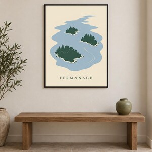 May include: A framed wall art print featuring a light blue lake with three dark green islands and a small white boat leaving a wake. The word FERMANAGH is printed in black capital letters at the bottom of the cream-colored background.
