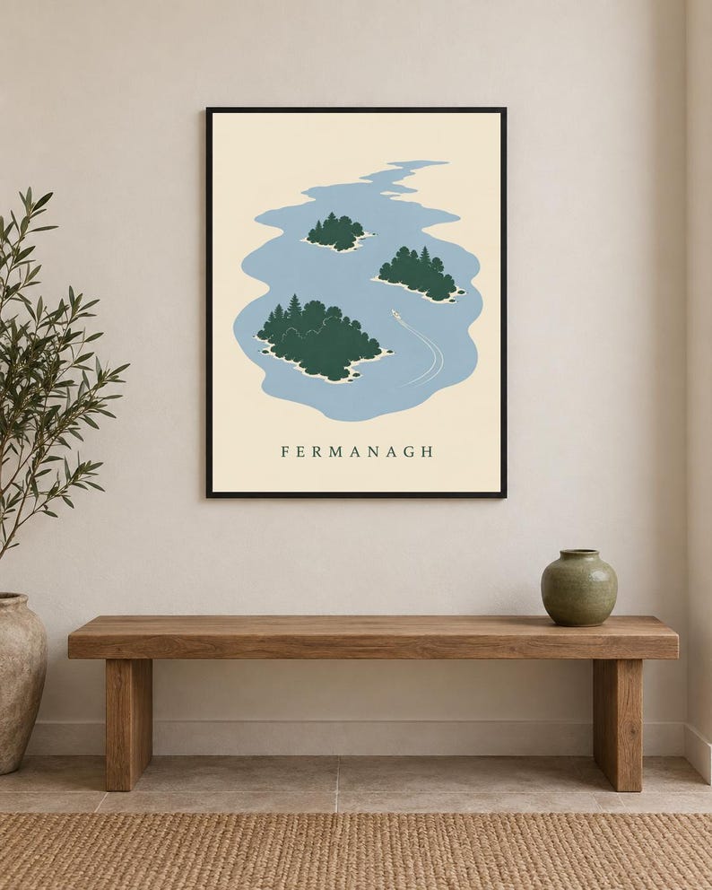 May include: A framed wall art print featuring a light blue lake with three dark green islands and a small white boat leaving a wake. The word FERMANAGH is printed in black capital letters at the bottom of the cream-colored background.