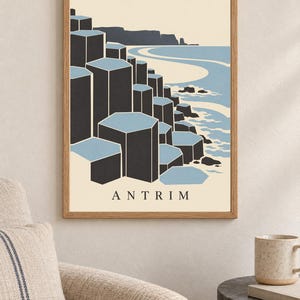 May include: A framed wall art print featuring a stylised illustration of the Giant's Causeway in Antrim. The graphic depicts dark grey hexagonal basalt columns against a light blue sea and cream sky, with the word ANTRIM printed in black at the bottom.