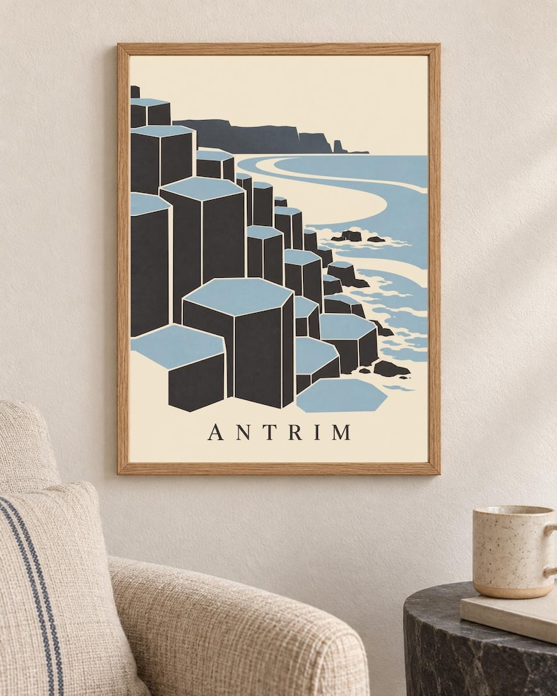 May include: A framed wall art print featuring a stylised illustration of the Giant's Causeway in Antrim. The graphic depicts dark grey hexagonal basalt columns against a light blue sea and cream sky, with the word ANTRIM printed in black at the bottom.