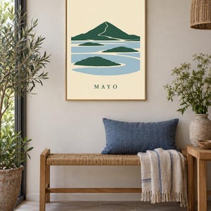 Pu&ograve; includere: Una stampa artistica da parete incorniciata che presenta un'illustrazione paesaggistica stilizzata di una montagna e isole circondate dall'acqua. Il design utilizza una tavolozza di verde scuro, azzurro e crema. La parola MAYO &egrave; stampata in basso.
