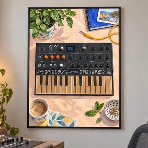 May include: A framed art print depicting an Arturia MicroFreak synthesizer with a black chassis, orange knobs, and a textured keyboard. The illustration includes a potted plant, a coffee cup, a small bowl with paperclips, and a blue notebook.