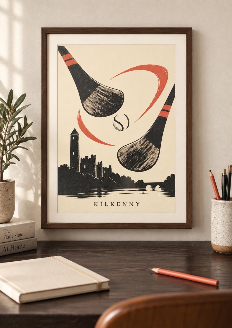 Peut inclure: Une impression d'art encadr&eacute;e illustrant deux crosses de hurling et une balle au-dessus de la silhouette du ch&acirc;teau de Kilkenny et d'une rivi&egrave;re. L'affiche est cr&egrave;me avec des d&eacute;tails noirs et rouges et le mot KILKENNY inscrit en bas.
