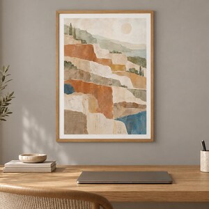 Op de afbeelding: Een ingelijste verticale wandkunstprint met een gelaagde landschapsillustratie, een bleke zon, terrasvormige heuvels in tinten terracotta, beige en saliegroen, en diepblauwe wateraccenten, gevat in een lichthouten lijst.