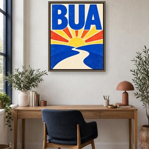 May include: A framed rectangular wall art print featuring the text BUA in large blue letters above a stylized yellow sun with red and yellow rays. Below the sun, a winding beige path leads through blue hills, creating a vibrant landscape scene.
