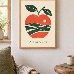 May include: A framed wall art print featuring a large red apple illustration on a cream background. The apple contains a landscape of green rolling hills and a small white sun. The word ARMAGH is printed in dark green capital letters below the apple.