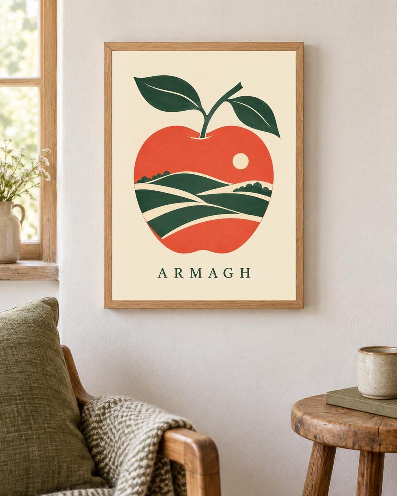 May include: A framed wall art print featuring a large red apple illustration on a cream background. The apple contains a landscape of green rolling hills and a small white sun. The word ARMAGH is printed in dark green capital letters below the apple.