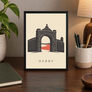 May include: A framed art print featuring a black graphic illustration of the historic stone city walls and arched gate of Derry. The design includes a red path leading through the archway against a cream background with the text DERRY below.