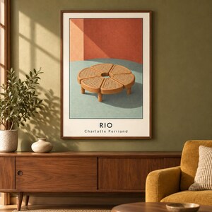 May include: A framed wall art print titled RIO by Charlotte Perriand, featuring a graphic illustration of a circular, segmented wooden coffee table with a woven cane surface set against a background of muted red and light blue geometric shapes.