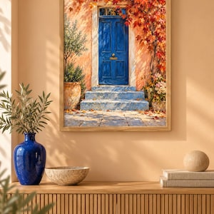 May include: A framed wall art print depicting a vibrant blue door set into a peach-coloured wall. The scene features stone steps, a potted olive tree, and climbing vines with autumn-toned red and orange leaves, all rendered in a painterly style.