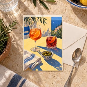 May include: A rectangular greeting card featuring an illustration of a yellow table set with an orange spritz in a stemmed glass, a dark red cocktail in a tumbler, a small bowl of green olives, and a white napkin with blue stripes.