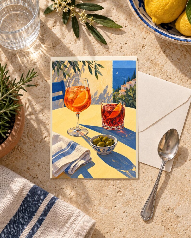 May include: A rectangular greeting card featuring an illustration of a yellow table set with an orange spritz in a stemmed glass, a dark red cocktail in a tumbler, a small bowl of green olives, and a white napkin with blue stripes.