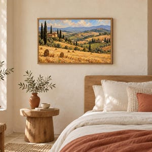May include: A rectangular landscape painting depicting a golden wheat field with hay bales, tall cypress trees, and rolling hills under a blue sky with clouds. The artwork is framed in a light wood finish and displayed on a textured off-white wall.
