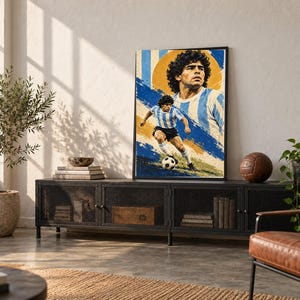 May include: A framed vertical poster featuring a stylized illustration of Diego Maradona in a light blue and white striped soccer jersey. The artwork displays a large portrait above a smaller action shot of the player running with a soccer ball.