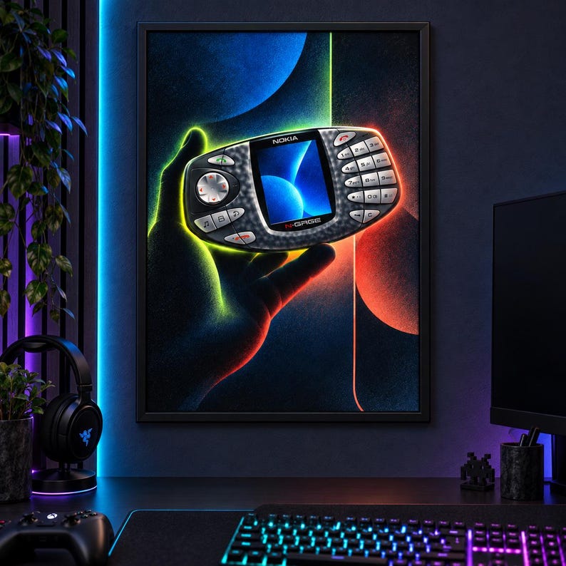 Puede incluir: Una impresi&oacute;n de arte de pared enmarcada que muestra un dispositivo de juego port&aacute;til Nokia N-Gage sostenido en una mano en silueta. El dispositivo es negro con una pantalla central que muestra gr&aacute;ficos azules, un pad direccional y un teclado.